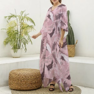Peut inclure: Une robe caftan longue jusqu'aux pieds avec une encolure en V et des manches courtes amples. Le vêtement est orné d'un imprimé intégral de plumes superposées dans des tons de rose poudré, mauve et blanc sur un fond rose clair.