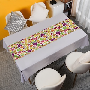 May include: A white tablecloth with a colorful fruit pattern table runner. The runner features a variety of fruits including cherries, bananas, pears, grapes, kiwi, and apricots. The tablecloth has a scalloped edge.