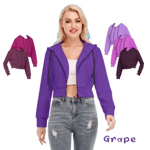 May include: A purple cropped zip-up hoodie with a hood and long sleeves. The hoodie has a front pocket and a black zipper. The model is wearing the hoodie with a white crop top and blue jeans. The text "Grape" is written in purple below the model.