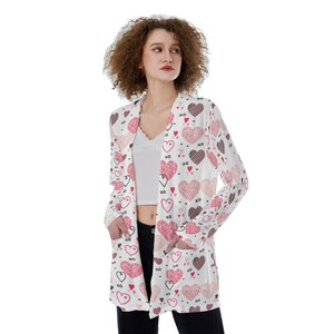 May include: A white cardigan with a long sleeve and a V-neck. The cardigan is covered in a pattern of pink and red hearts with the letters XO printed on them.