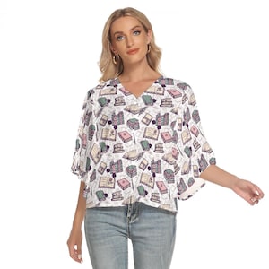 May include: White blouse with a v-neck and short sleeves. The blouse features a print of books and flowers.