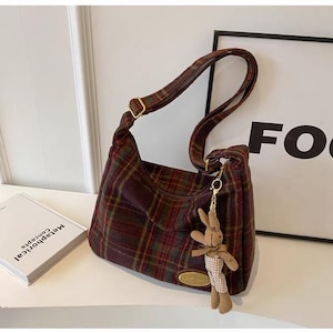 May include: A hobo-style shoulder bag featuring a dark red, green, and yellow plaid pattern. The bag includes an adjustable strap with gold-tone hardware, a small oval gold-tone brand plaque, and a plush brown rabbit charm attached to the side.