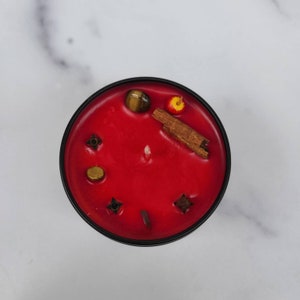 May include: A red candle with embedded crystals and spices, including cinnamon sticks, cloves, and tiger's eye.