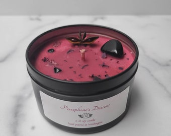 May include: A 6 oz soy candle in a black tin with a pink wax base. The candle is decorated with dried flowers, black stones, and a star anise. The label reads "Persephone's Descent 6 oz soy candle hand poured in Washington."