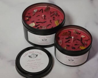 May include: Two black metal candle tins with red wax and embedded crystals. The labels read "Kitchen Witch" and "hand poured in Washington". The smaller tin has a lid that reads "Violet Moon Candles Kitchen Witch" with a description of the candle's scent and properties.