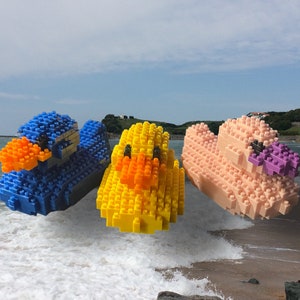 May include: Three toy ducks made of colorful plastic building blocks. The ducks are blue, yellow, and pink. They are floating in the air above a beach with waves crashing on the shore.