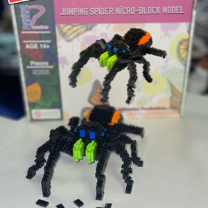 May include: A black and orange jumping spider micro-block model with green eyes and blue accents. The model is assembled and sitting on a white surface. There are extra black micro-blocks scattered around the model. The box for the model is in the background and says "Jumping Spider Micro-Block Model" on the front.