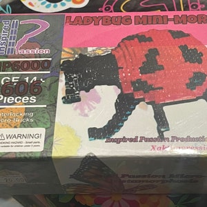 May include: A box for a ladybug mini-morph 3D puzzle. The box is pink and white with a picture of a red and black ladybug on the front. The box says "LADYBUG MINI-MORPH" and "IP6000". It also says "AGE 14+ 606 Pieces" and "Interlocking Micro Bricks".