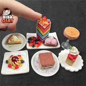 Può includere: Diversi dessert in miniatura, tra cui una torta arcobaleno e tiramisù, presentati su piccoli piatti bianchi. L'immagine presenta anche una ciambella su un supporto di vetro, bacche e gelato. Il logo "Hiba Clay" è visibile.