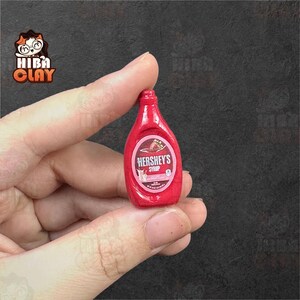 May include: Miniature red Hershey's syrup bottle. The bottle has a white and pink label with the Hershey's logo and a strawberry illustration. The bottle is held between fingers against a dark background.