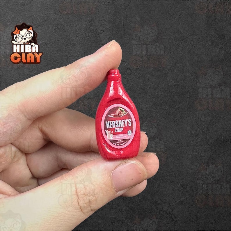 May include: Miniature red Hershey's syrup bottle. The bottle has a white and pink label with the Hershey's logo and a strawberry illustration. The bottle is held between fingers against a dark background.