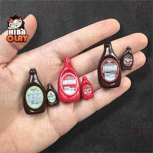 May include: Miniature Hershey's syrup and ketchup bottles in various sizes. The bottles are brown and red, with labels. The image shows a collection of small, detailed food replicas, perfect for dollhouses or crafts.