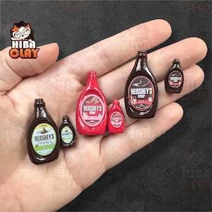 May include: Miniature Hershey's syrup bottles in various sizes, including red and brown bottles. The bottles have the Hershey's logo and text. The bottles are held in a hand.