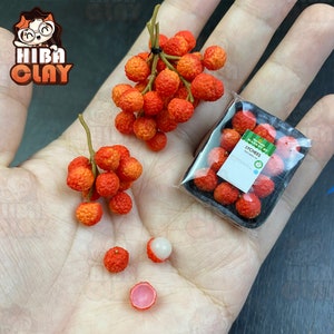 May include: A close-up of a hand holding three bunches of miniature lychee fruit. The lychees are red and have a rough texture. One lychee is cut open, revealing a white flesh. The lychees are arranged in a plastic container with a label that reads "LYCHEES".
