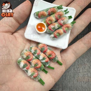 May include: Miniature food set featuring spring rolls with shrimp and a small dipping sauce on a white plate. The spring rolls are wrapped in a translucent material, showcasing the shrimp and green herbs inside. The set is designed to look like a realistic meal.