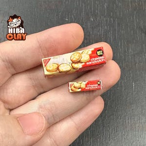 May include: Miniature boxes of Ritz cheese sandwich crackers. The boxes are red and white with images of the crackers. The text "Ritz cheese sandwich" is visible on the boxes. The boxes are held in a person's hand.