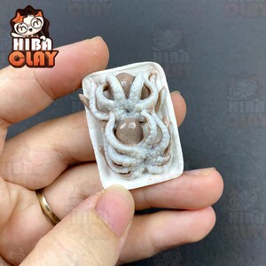 May include: A miniature white ceramic dish with a brown octopus inside. The octopus has white tentacles and is arranged in a circular pattern. The dish is held in a person's hand.