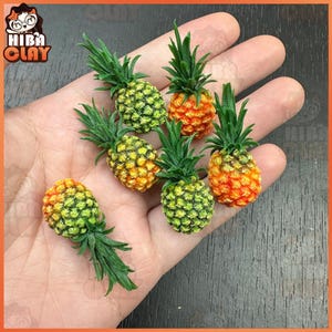 Op de afbeelding: Een hand die zes miniatuur-ananas vasthoudt. De ananas zijn gemaakt van polymer clay en hebben een realistische uitstraling. Ze zijn geschilderd in tinten groen, geel en oranje.