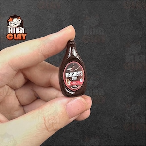 May include: Miniature Hershey's Syrup bottle. The dark brown bottle has a white and red label with the text "HERSHEY'S SYRUP". The bottle is held in a person's hand. The background is a dark gray color.