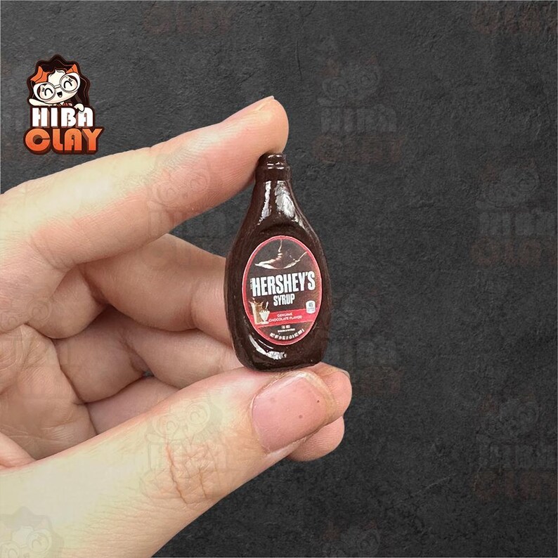 May include: Miniature Hershey's Syrup bottle. The dark brown bottle has a white and red label with the text "HERSHEY'S SYRUP". The bottle is held in a person's hand. The background is a dark gray color.