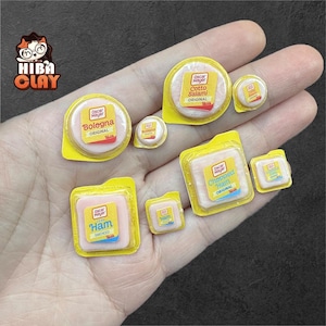 May include: Miniature food items including bologna, cotto salami, and ham, each in individual yellow packaging. The labels read "Oscar Mayer" and product names. The items are arranged on a hand, showcasing the small scale of the food replicas.