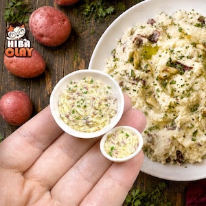 May include: Two miniature white bowls filled with a textured, creamy mashed potato mixture containing visible red potato skins and green herbs. The bowls are held in a palm, positioned next to a larger plate of the same mashed potato dish.