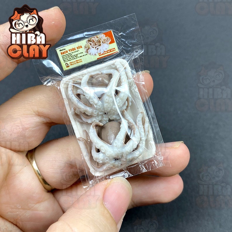 May include: A clear plastic bag containing a small white styrofoam container with two small, cooked octopus. The container has a label that reads "Bạch Tuộc Sữa" in Vietnamese.