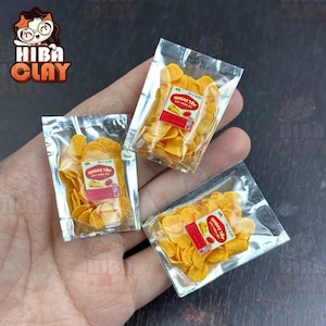 May include: Miniature bags of potato crisps. Each clear plastic bag is filled with golden-yellow crisps and a red and white label. The bags are arranged on a surface. The logo "Hiba Clay" is in the upper left corner.