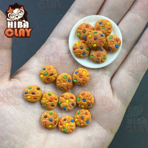 Op de afbeelding: Een hand die een bord met kleurrijke chocolade-chipkoekjes vasthoudt. De koekjes liggen op een wit bord en worden omringd door meer koekjes op de hand.