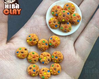 May include: A hand holding a plate of colorful chocolate chip cookies. The cookies are on a white plate and are surrounded by more cookies on the hand.