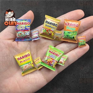 Puede incluir: Pequeños paquetes de caramelos Haribo en varios sabores, incluyendo Sour Sghetti, Goldbears, Frogs, Peaches y Twin Snakes. Las coloridas bolsas se sostienen en una mano, mostrando el diseño detallado de cada paquete.