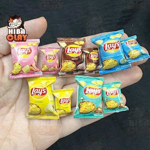 Könnte beinhalten: Eine Sammlung von Miniatur-Lay's-Kartoffelchip-Tüten in verschiedenen Geschmacksrichtungen, darunter Rosa, Braun, Gelb und Blau. Die Tüten zeigen das Lay's-Logo und Bilder von Chips. Das Bild zeigt eine Hand, die die Miniaturtüten hält.