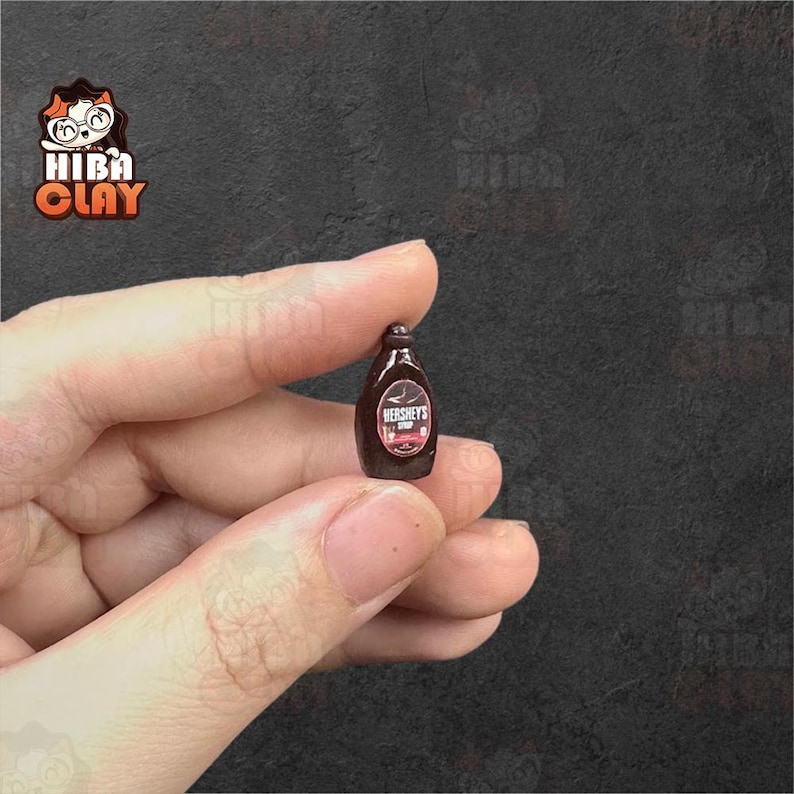 May include: Miniature Hershey's syrup bottle. The brown bottle has a white and red label with the Hershey's logo. The bottle is held between two fingers against a dark background. The bottle is a small, detailed replica.