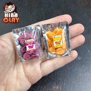 May include: Two small bags of dried sweet potato chips. The bag on the left contains purple sweet potato chips and the bag on the right contains orange sweet potato chips. Both bags have labels with Vietnamese text.