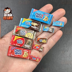 Pode incluir: Pequenas embalagens de biscoitos Chips Ahoy! em vários tamanhos e designs, seguradas em uma mão. As embalagens apresentam o logotipo Chips Ahoy! e a marca Hershey's, com embalagens vermelhas, azuis e marrons.