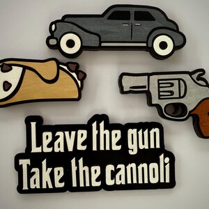 May include: Wooden cutouts of a gray vintage car, a cannoli, a silver revolver, and a black sign with the text "Leave the gun Take the cannoli" in white. The objects are on a white background.