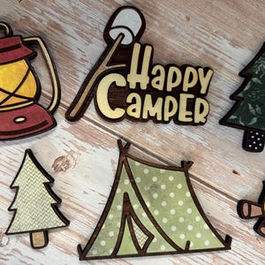 May include: A collection of wooden camping-themed cutouts. The set includes a lantern, a marshmallow on a stick, the words "Happy Camper", two trees, a tent, and a campfire. The cutouts are in various colors, including red, yellow, green, and brown.
