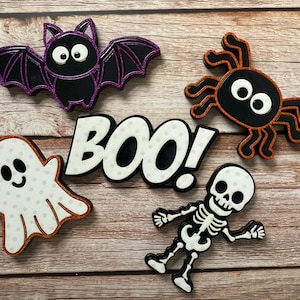 Puede incluir: Decoraciones temáticas de Halloween que incluyen un murciélago negro con alas moradas, una araña negra con detalles naranjas, un fantasma blanco con lunares, un esqueleto y la palabra "BOO!". Las decoraciones están sobre una superficie de madera.