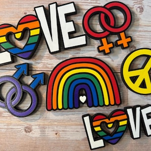 Peut inclure: Divers éléments décoratifs en bois sur le thème de l'arc-en-ciel, dont le mot "LOVE", un arc-en-ciel, des cœurs, un symbole de paix et des symboles de genre. Les lettres sont blanches avec des bordures noires, et les cœurs et l'arc-en-ciel présentent des couleurs vives.