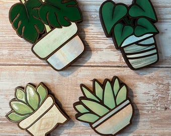 May include: Four wooden plant-themed decorations. Each features a different plant design in a pot. The plants are green, and the pots have a light color scheme with brown edges. The designs include a monstera, a heart-leaf philodendron, and two succulent plants.