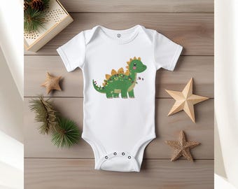May include: A white short-sleeve baby bodysuit featuring a green dinosaur graphic with orange spikes and colorful string lights. The text 5 GRÖBEN 5 SIZES is at the top, and DST, EXP, HUS, JEF, PES, VIP, VP3, XXX are printed at the bottom of the image.
