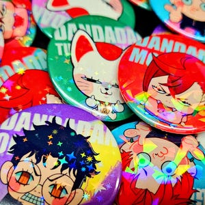 以下が含まれることがあります： 様々な漫画のキャラクターと「Jandada」という文字が書かれた、カラフルでホログラフィックなボタンのコレクション。ボタンは星とグリッターで飾られています。