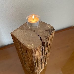 Puede incluir: Un portavelas rústico de madera con una vela de té encendida. La vela se encuentra sobre un trozo de madera cortado en forma cuadrada, mostrando la veta y textura natural. La madera es de color marrón cálido y la llama de la vela brilla intensamente.
