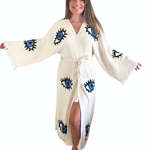May include: A white cotton robe with blue and black eye designs. The robe has a long sleeve and a wrap style closure. The robe has a fringe trim.