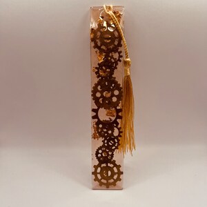 May include: A gold bookmark with a tassel, featuring a design of gears and gold leaf embedded in clear resin.