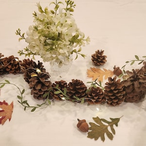 May include: A garland of pine cones and greenery with white lights, accented with fall leaves and acorns. The garland is on a white tablecloth with a bouquet of white flowers in a clear vase.