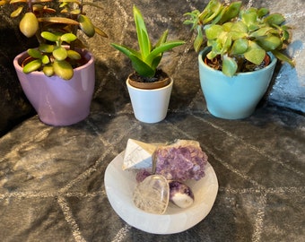 May include: A white selenite bowl with a purple amethyst cluster, a white selenite pyramid, and a clear quartz heart crystal. The bowl is sitting on a gray and white patterned surface.