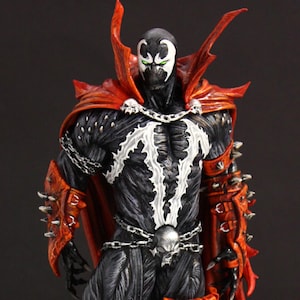 May include: An action figure of Spawn, a comic book character, standing on a green and grey base. The figure is wearing a red cape and has a skull on its belt.