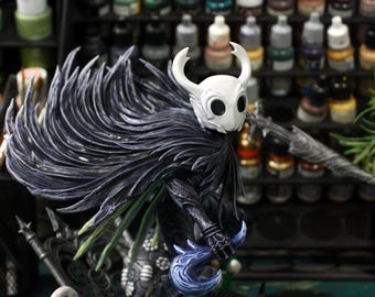 Puede incluir: Figura detallada de un personaje con máscara blanca y cuernos curvos, que lleva una capa oscura. La figura está posada sobre una base rocosa con una hoja azul. El fondo muestra botes de pintura en un estante.