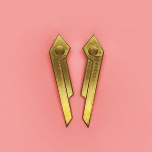 May include: Two gold-colored earrings with a geometric design. Each earring has a diamond-shaped top with a circular detail and a long, rectangular body. The earrings are set against a pink background.